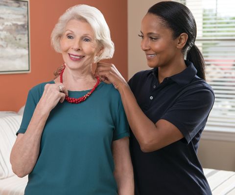 A cheery Erickson employee assists a smiling resident with putting on her necklace. 